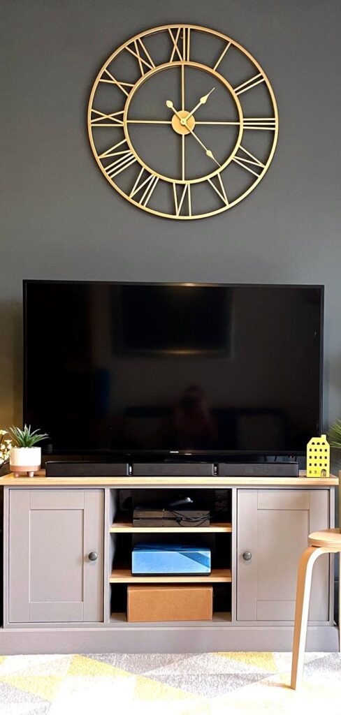 A large gold clock on a grey wall with a television on a cabinet